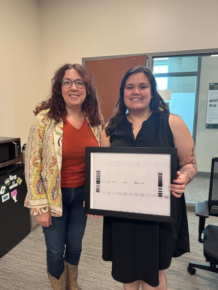 Two women smiling. One is holding a framed gel electrophoresis image