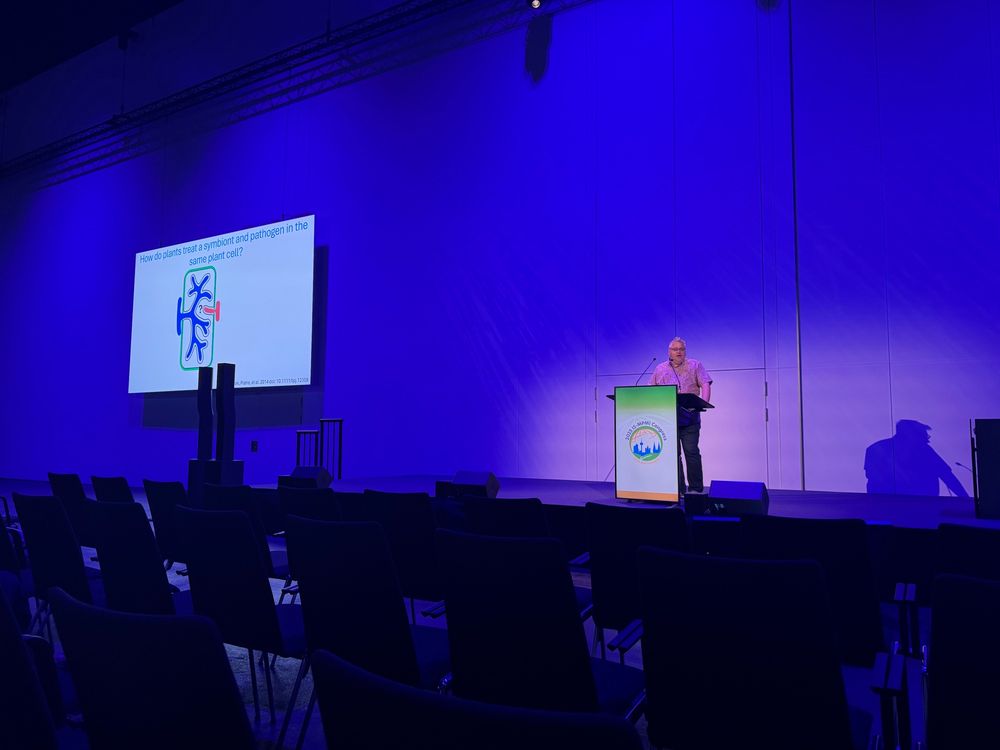 A person giving a talk on a stage with purple lighting. A slide asks the question how do plants treat a symbiont and pathogen in the same cell?
