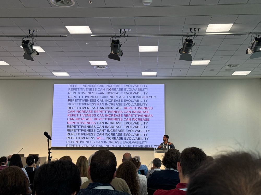 A person presenting with a PowerPoint slide that has repetitiveness can increase evolvability written over and over with slight changes indicated in red, such as changing from can to will