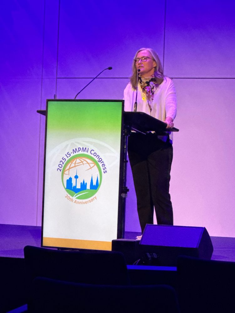 A woman speaking at a lectern with the MPMI logo