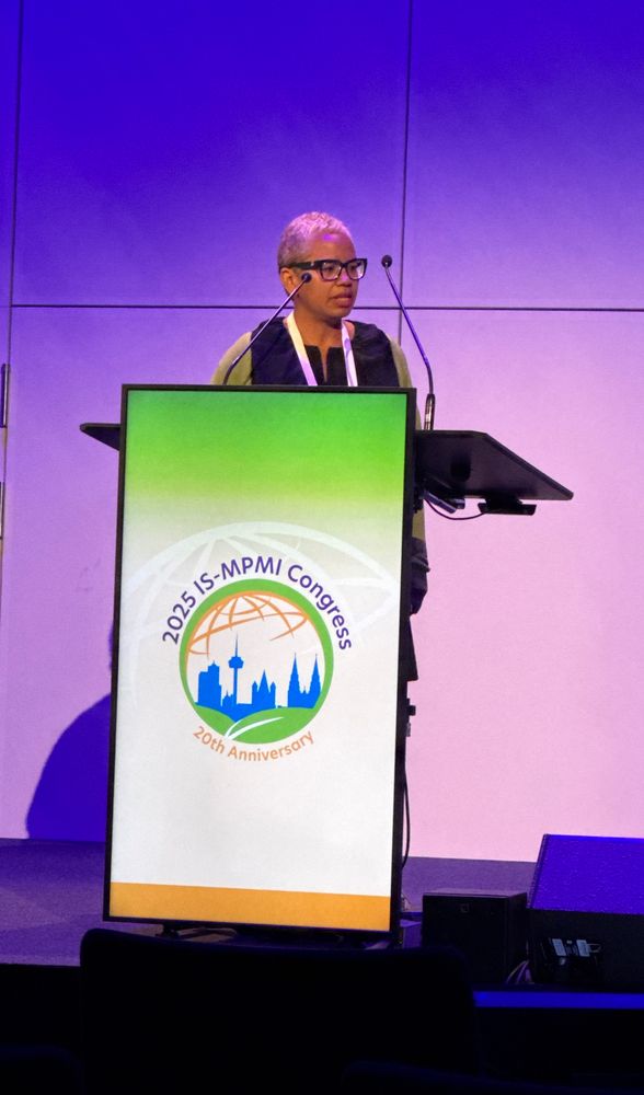 A woman speaking at a lectern with the MPMI logo