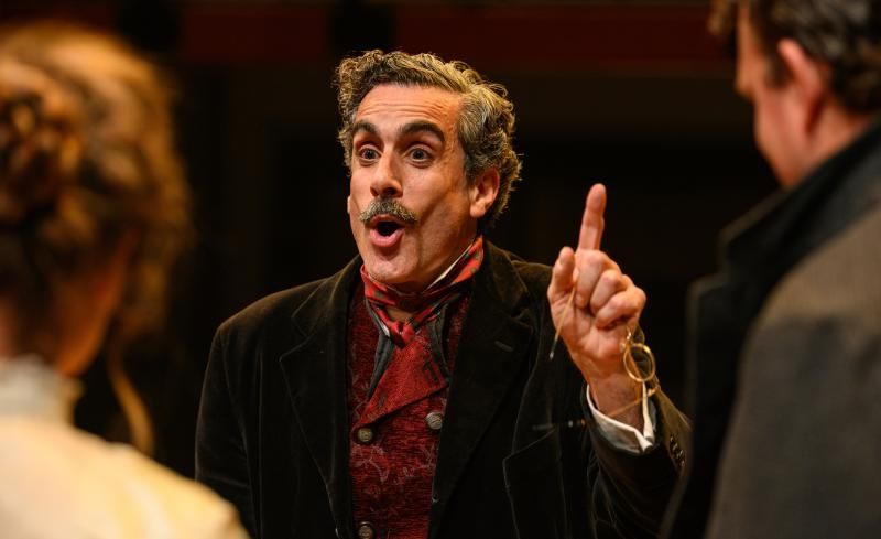 We're looking over the shoulders of Leoncavallo (right) and his wife (left) to a head and shoulders view of Torkia as Puccini, in the middle of saying something to them. He has a gleeful expression and is holding up his left index finger as if making a point. He wears a moustache, dark jacket and dark red silk cravat 