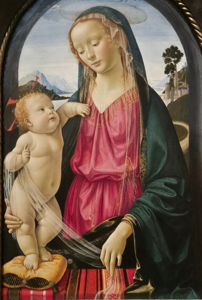 Madonna and Child. The Virgin stands in an arched window, wearing a red dress and dark hooded cloak, with a fine veil just visible under it. She's looking down  tenderly at the infant Jesus. He stands on her right, on a gold cushion on the sill in front of her. This also has a richly-coloured striped cloth over it. Jesus is looking back up at his mother.  He rests his left arm on her shoulder and holds on to  her dress with his left hand. He holds up a finger of his right hand as though he's preaching. Another veil is looped over the crook of his right elbow and she's holding the end of it with her left hand, while her right hand supports his chubby leg. The backs of their heads, the veil and the Virgin's left arm make an oval framing their faces. There's blue sky, hills and a lake behind them. 