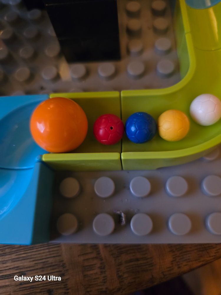 Photo of several lego & a Duplo ball sitting in a marble run track. The lego balls are significantly smaller & feel like they weigh more..
