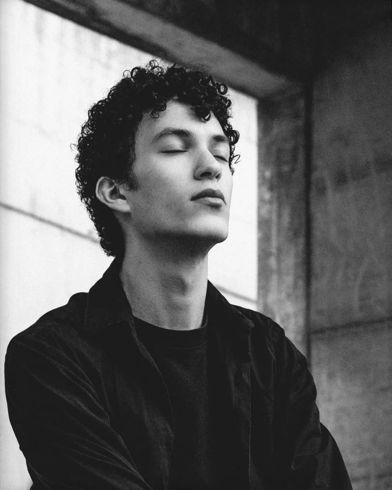 monochrome portrait of a young man wearing a black outfit while sitting in a urban area build out of nothing but concrete
