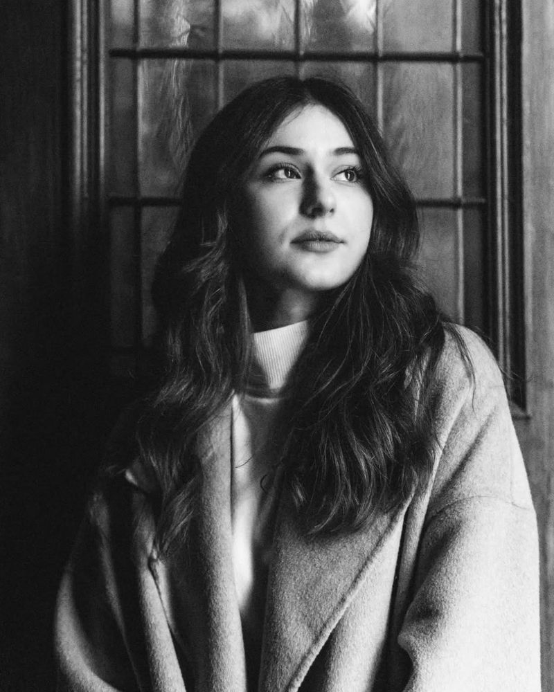 black and white photo of a young woman with long dark hair, wearing a bright coat. She looks to the side into the light, behind her is a old wooden door with a glass window