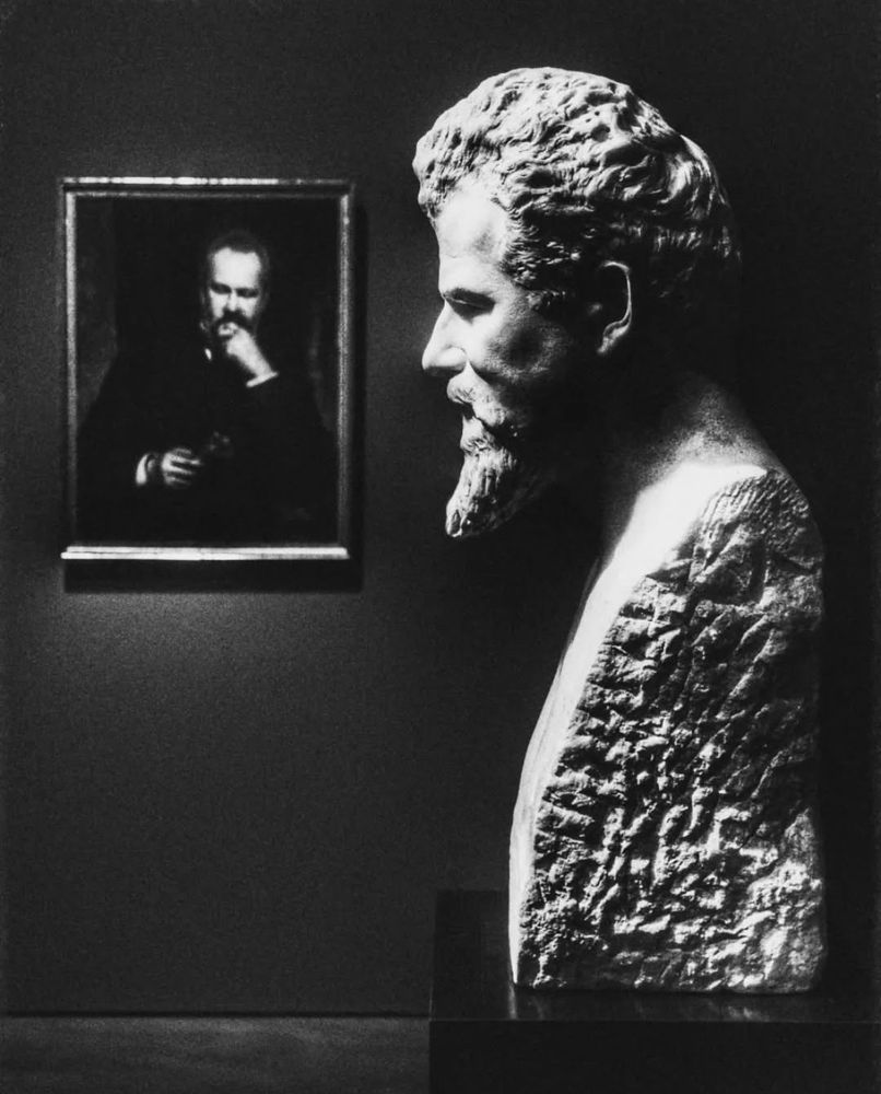 black and white photograph showing a old stone bust and in the background a painted portrait of a man