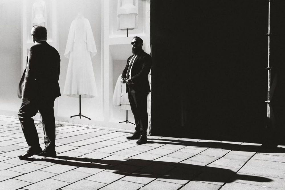 black and white street photo of a security guard in front of a shop. another older man walks by and casts a long shadow on the ground