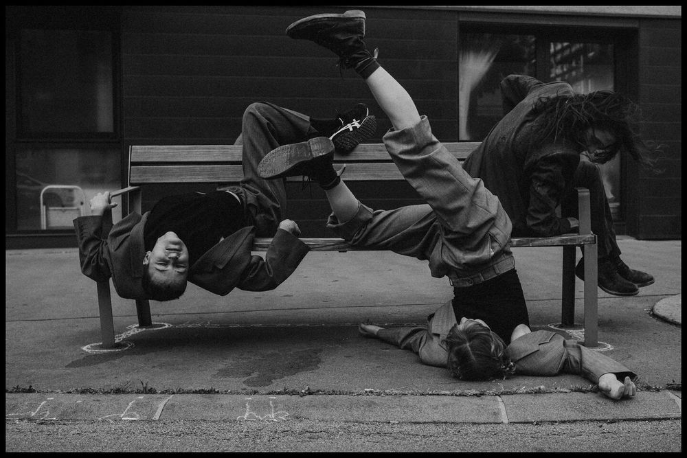 three young people wearing suits using a bench next to the street in unusual ways 