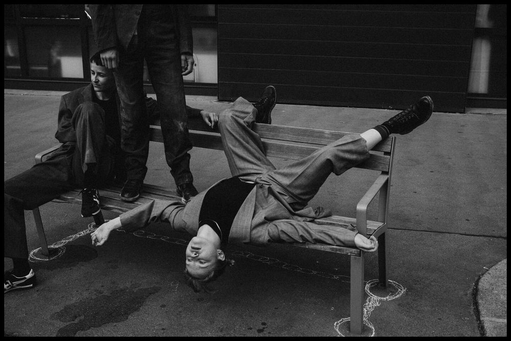 three young people wearing suits using a bench next to the street in unusual ways 