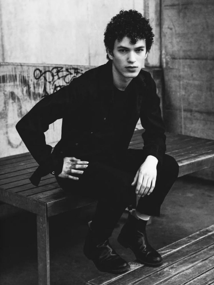 monochrome portrait of a young man wearing a black outfit while sitting in a urban area build out of nothing but concrete