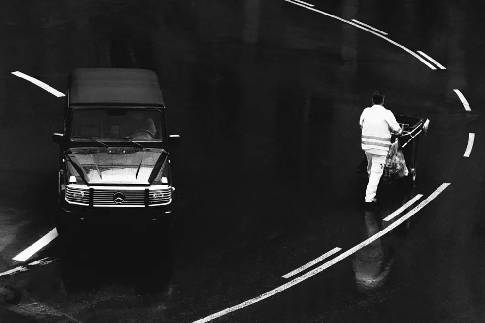 monochrome street photo of an black Mercedes G-Class SUV next to a worker pushing a garbage can