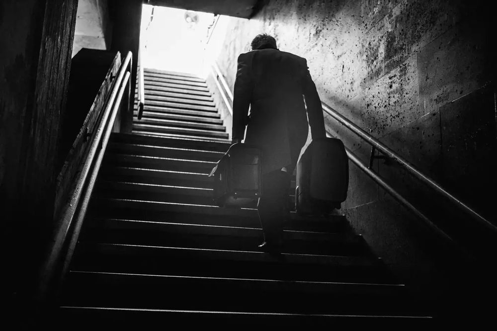 dark black and white photograph of a man wearing a short coat. He is climbing the stairs of a subway station and is carrying 2 heavy looking bags