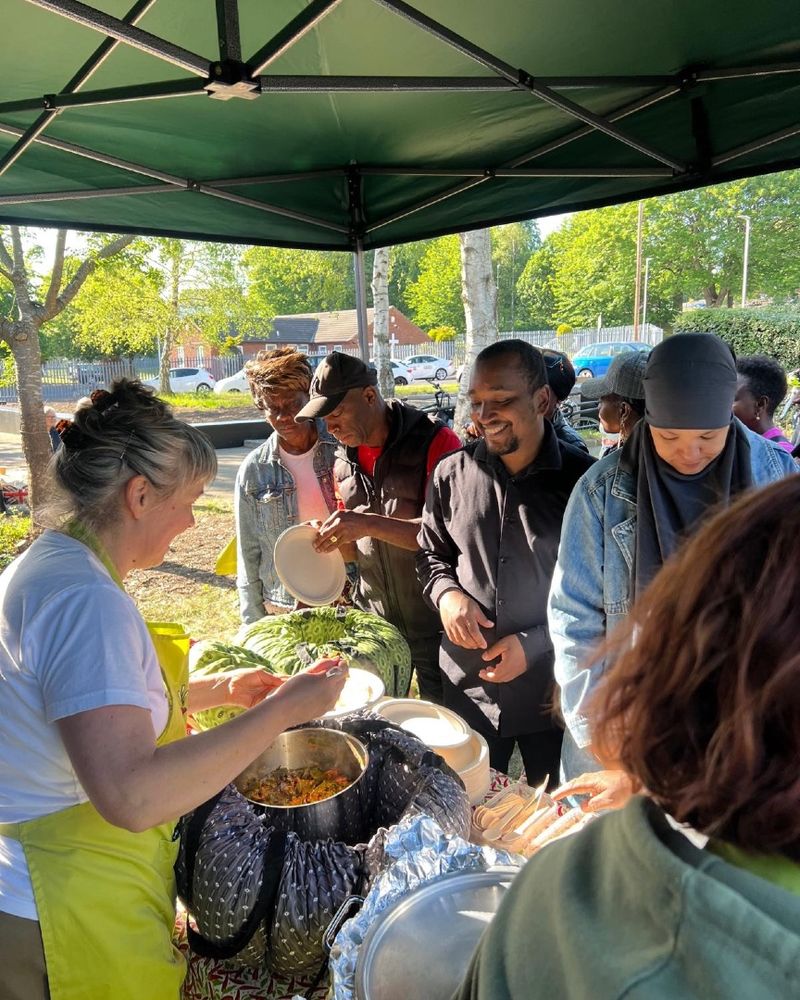 2 women serving food under a canopy - people of different skin colours including black, brown and white
