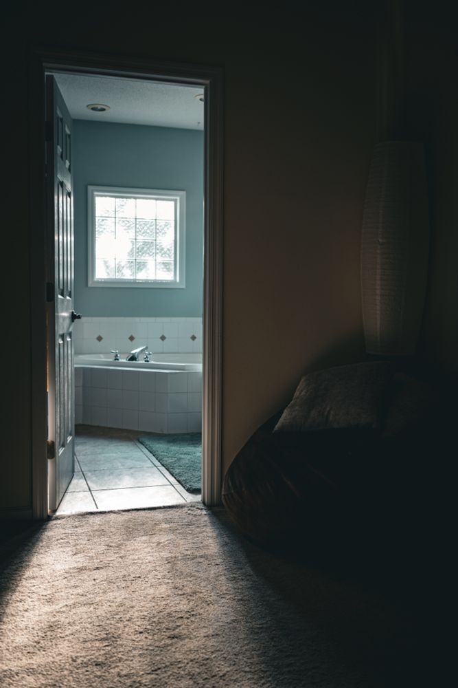Partially open door leading from a carpeted room into a bathroom with tiled floors. Sunlight streams through a frosted glass window, casting light onto a white bathtub with chrome faucets. A green bath mat lies on the floor near the tub. In the darker room outside the bathroom, a large beanbag with a pillow is visible, along with a tall floor lamp against the wall.