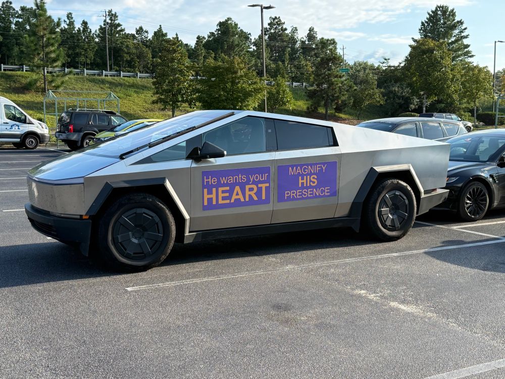 A Tesla Cybertruck is parked with a large purple decal on its side that reads “He wants your HEART” and “MAGNIFY HIS PRESENCE.” Other cars and trees are visible in the background.