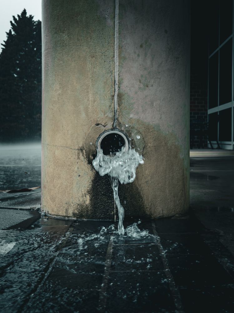 Water flows forcefully from a circular drain embedded in a large, weather-stained concrete pillar. The water splashes onto dark, wet tiles below and spreads outward, creating ripples and reflections. The surrounding area appears damp and overcast, with rain in the background and a partially visible glass building to the right. A tall, dark tree stands on the left side in the distance.