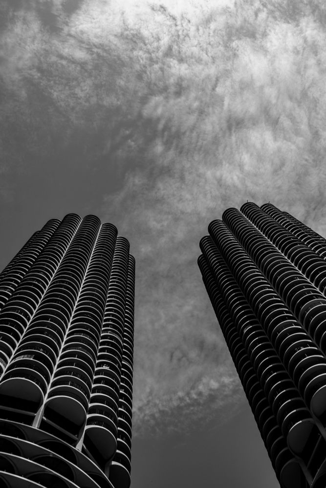 Two tall cylindrical skyscrapers with repeating curved balcony patterns rise against a partly cloudy sky, photographed from a low upward angle in black and white. The towers stand parallel to each other with a gap of open sky between them, creating a symmetrical composition.