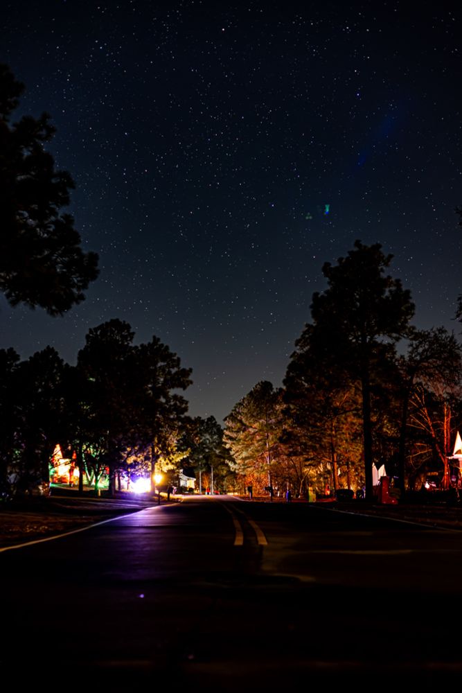 A nighttime scene featuring a paved road with yellow centerlines, surrounded by tall trees illuminated by artificial lights. The trees on the right display warm tones of orange and red, while those on the left are lit with colorful lights, including green and red. The sky is clear and filled with numerous stars, with a faint blue glow visible near the upper right. The road is slightly reflective, and scattered light sources illuminate parts of the scene, creating a contrast against the dark environment.