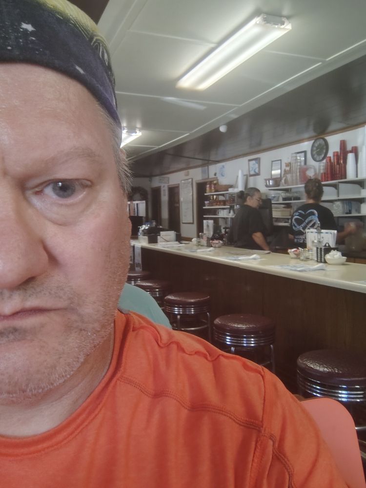 Half the face of Middle aged man (me) discreetly photographing the scene behind me, an old school diner bar with stools and two ladies behind the counter. Fluorescent lighting, no frills, perfect. 