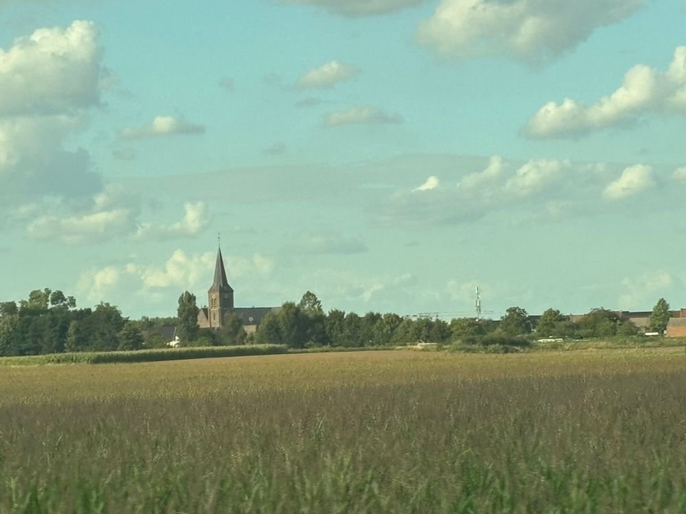 Church spire in a flat landscape under a cloud-dotted sky. The words are from Jacques Brel’s song « Le Plat Pays » about just this sort of landscape. 