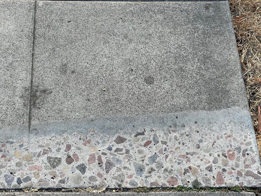 A sidewalk tile where the top layer of plain grey concrete has been scraped away along the bottom edge, revealing colorful pebbles inside the concrete, giving it a terrazzo-like appearance.
