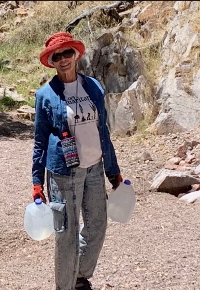 My beautiful, 85 year old mom carrying two jugs of water in the Arizona Sonoran desert. She’s wearing long pants, long sleeves, and a hat to protect her pale Norwegian skin from the sun. Her shirt says Los Samaritanos (Samaritans). 

She’s smiling. 
She’s almost always smiling. 