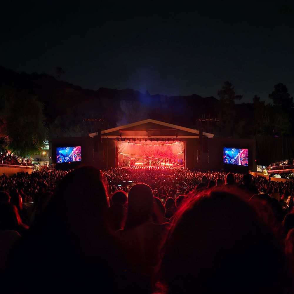View of the Greek Theater stage, lit orange-red, screens on either side and a sea of people in between