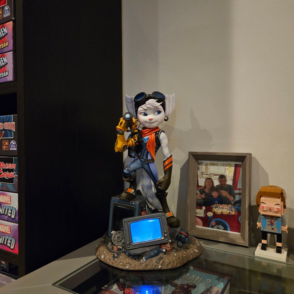 The restored Rivet statue, with the TV screen in the base lit up blue, sits on top of a grey-framed glass shelf, between family pictures and stacks of board games.