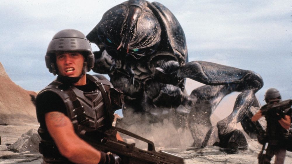 Still from classic satire of fascism, Starship Troopers: a massive, black beetle climbs up through the ground behind two fleeing soldiers. One further away and to the right has their face obscured by their rifle, the closer one is facing toward us clearly has the face of central character Johnny Rico