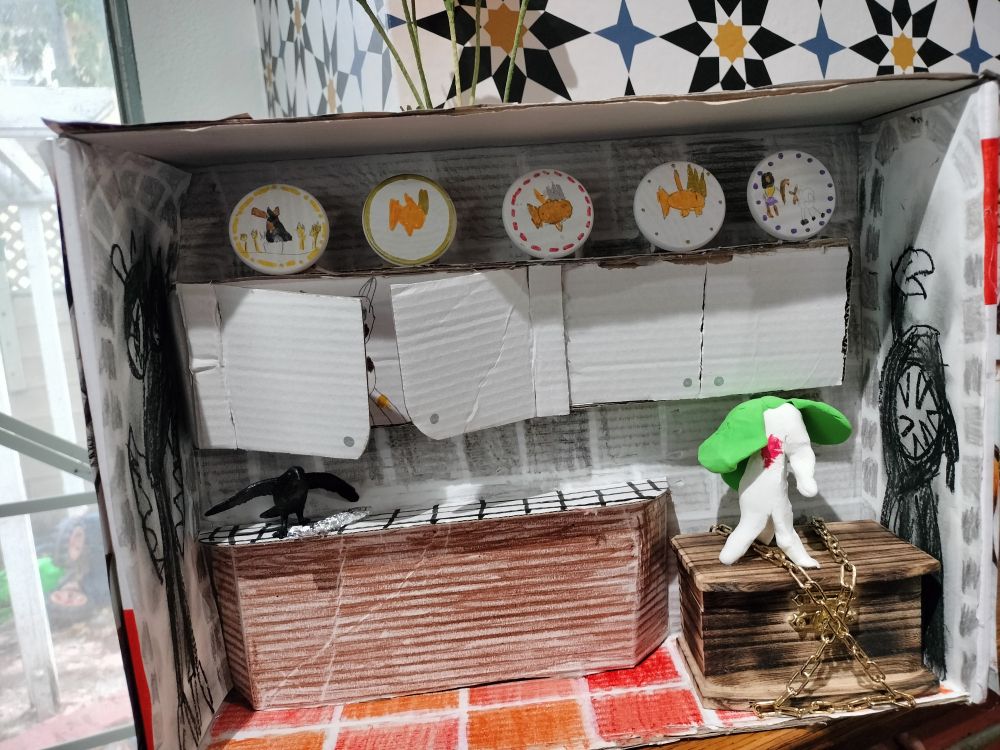 A child's book report diorama in a small cardboard box. It depicts a kitchen scene with cabinets made of cardboard and tiled walls done in crayon. The top of the cabinets has a row of decorative plates and there is a large box in the corner of the floor with chains wrapped around it. A white, carrot-shaped creature stands atop the box. The Scarling from Illuminations by T. Kingfisher. On the kitchen counter a crow stands over a shiny spoon made of aluminum foil.