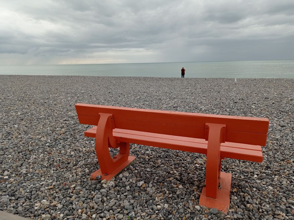 Un banc orange tourné  vers l'océan sur une plage de galets. On aperçoit une personne debout proche de l'eau