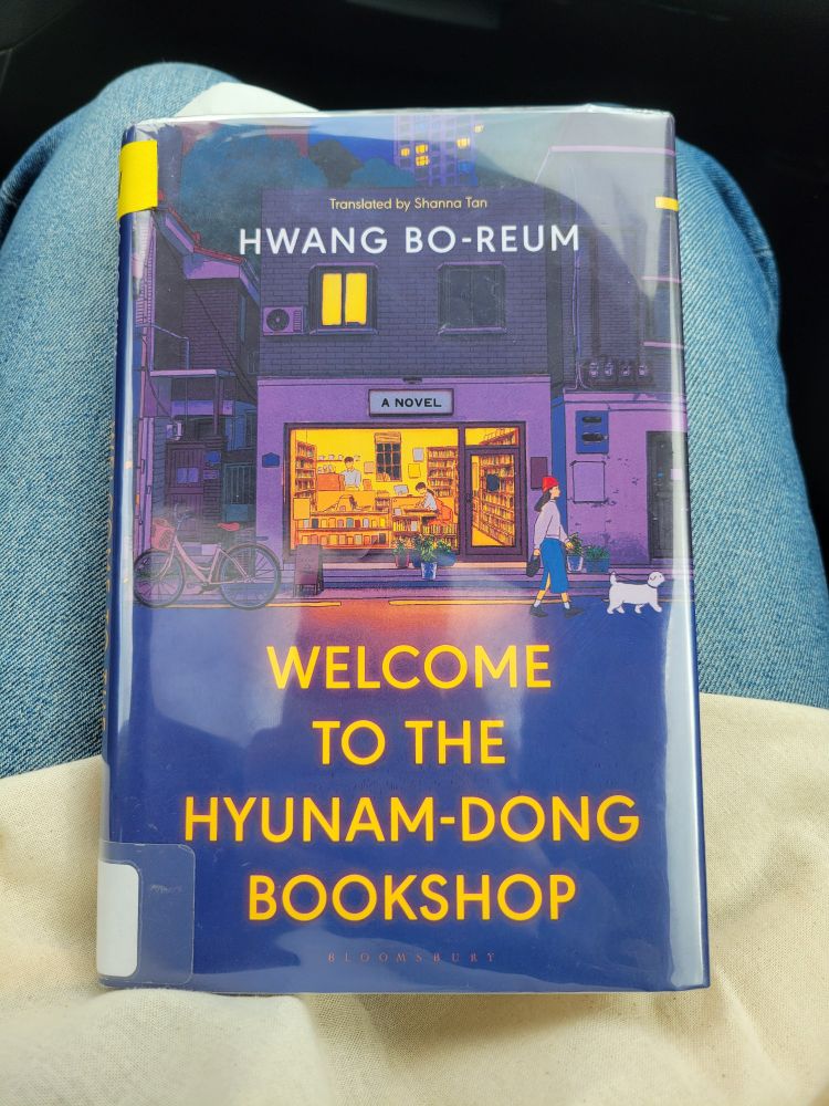 Book cover for Welcome to the Hyunam-Dong Bookshop by Hwang Bo-Reum (trans. Shanna Tan). Exterior of a bookstore at night. Inside a man stands behind a coffee counter and a woman sits writing. Outside, a woman walks by with a small white dog. 