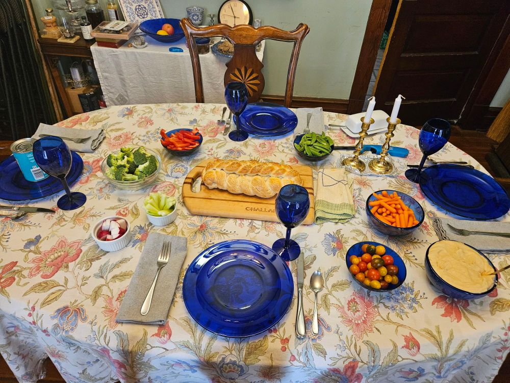 A table with a challah in the center, a large bowl of homemade hummus, and smaller bowls with Radishes, broccoli, red peppers, sugar peas, carrots, and small tomatoes. 
It was so hot today that the menu shifted last minute to accommodate lighter foods.