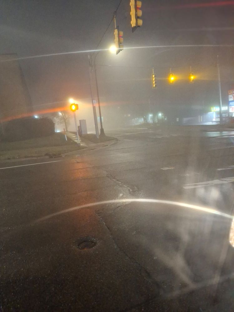 A picture of the local streets at nighttime. Unfortunately the weather decided to be so foggy as to make it hard to see down the road. Image taken from inside my car, at a red light.