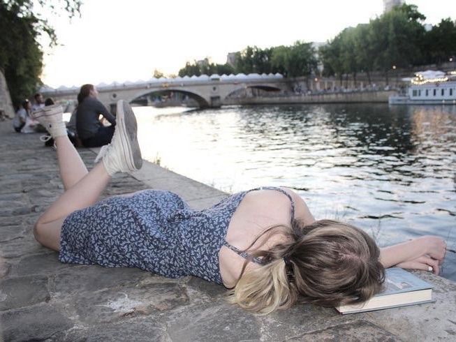 A girl wearing white Converse and a blue sun dress is lying on the grounf by a canal or a river with her head on a book.
