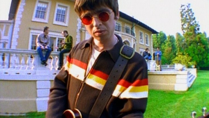 A Gallagher of some sort from Oasis in a video screen shot where he's in a snappy brown and orange striped 1970s retro shirt.