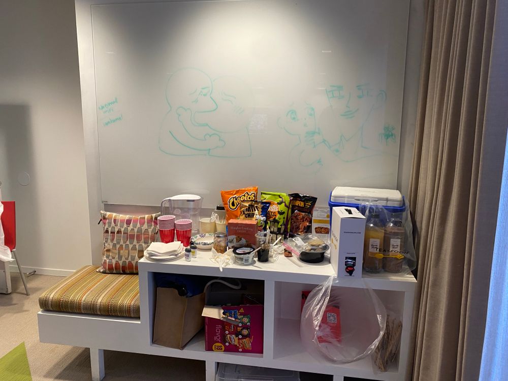 display of an array of food/snacks with whiteboard having two yaoi-related memes on them, one of them of two characters kissing and the other depicting caricatures of an uke and seme