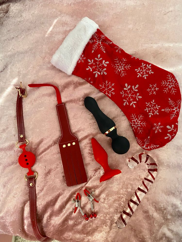 A red Christmas stocking with snowflakes next to a red ball gag, red paddle, red jingle bell nipple clamps, a red butt plug, a black vibrator, and a glass candy cane dildo 