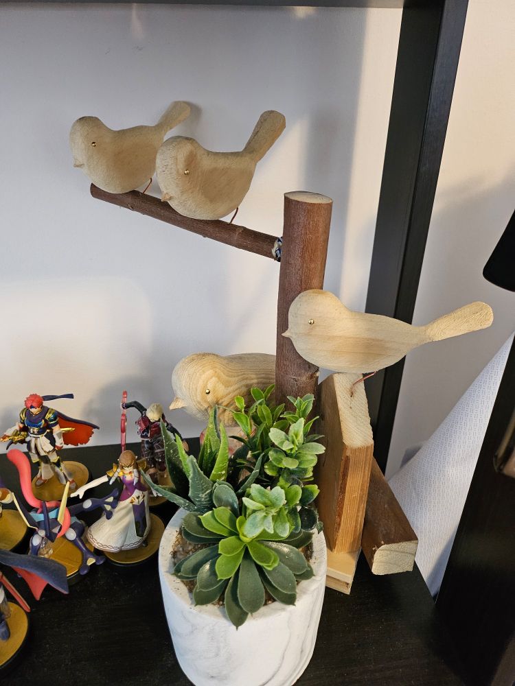 Hand carved woodwork of birds on a branch, unpainted. Some amiibos and a fake plant are placed nearby.