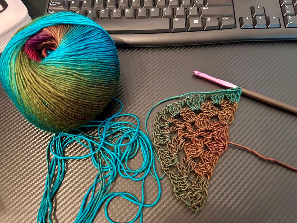 A ball of yarn showing shades of blue, green, & brown on a black table. The start of a crochet project in a triangle shape with a pink and black crochet hook next to it.  

Yarn is Mary Maxim Prism in the color Autumn Mist. 