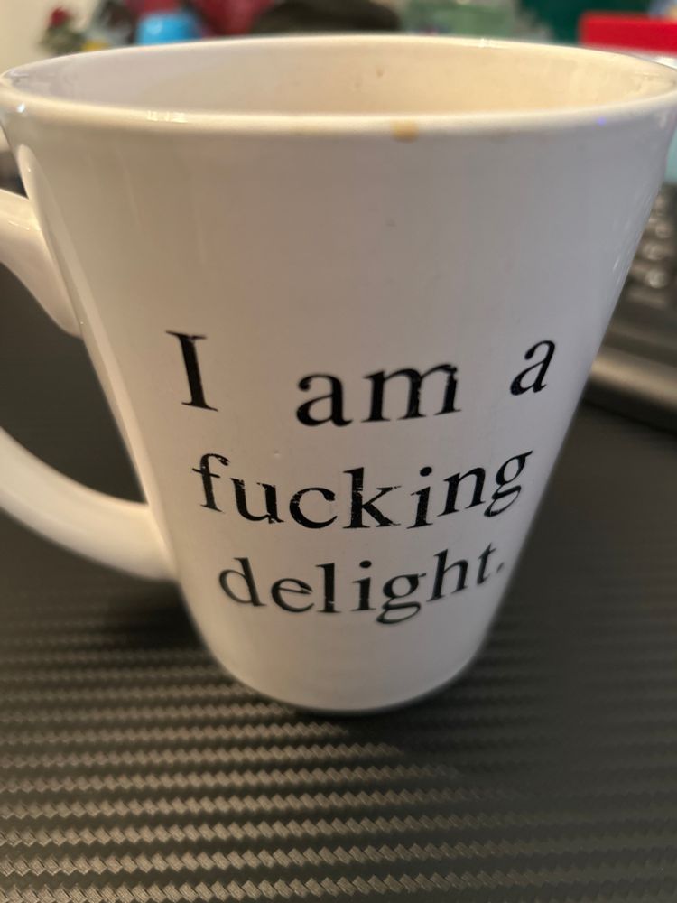 White coffee mug on a black desk. The words are the mug are, “I am a fucking delight.” 
