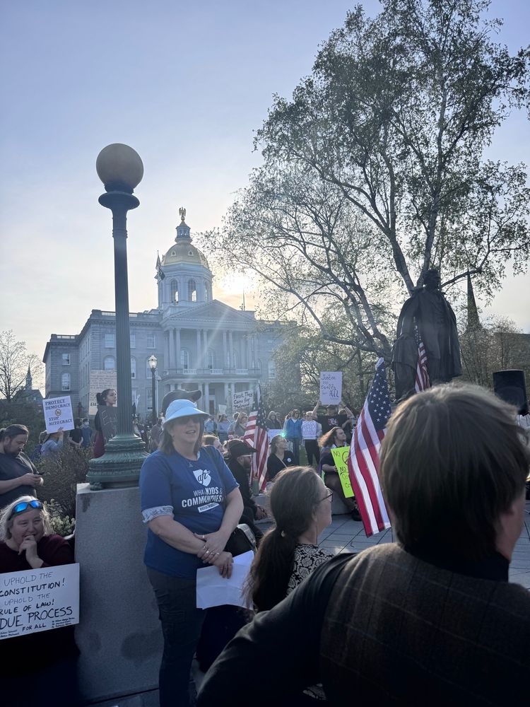 The sun sets behind the Concord State House. People in the foreground. Signs and flags. 