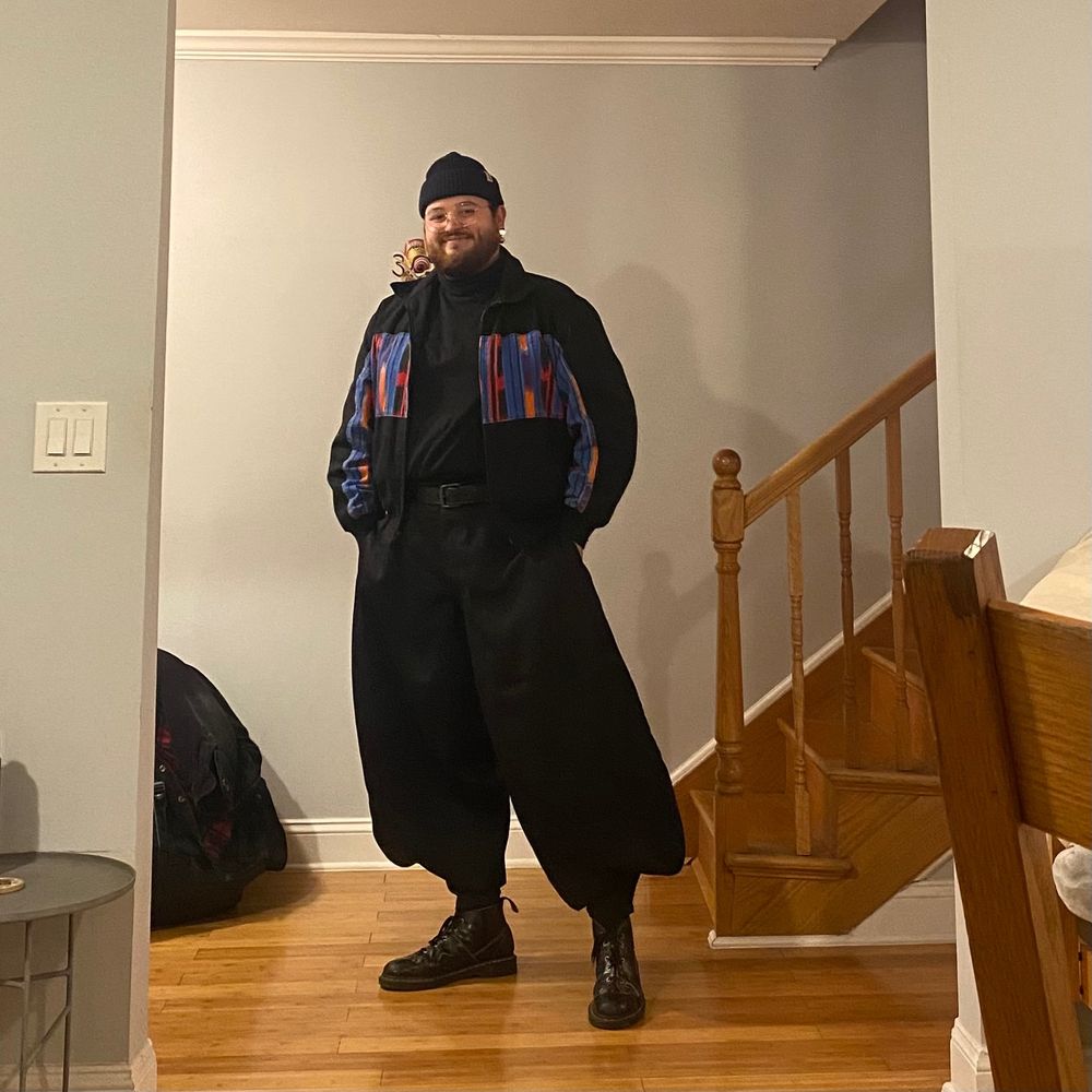 bearded person standing in front of a staircase wearing a pair of black, Tobi-style balloon pants, black glossy boots, and a black cropped jacket with a red, blue, red and orange abstract pattern on the chest of the jacket
