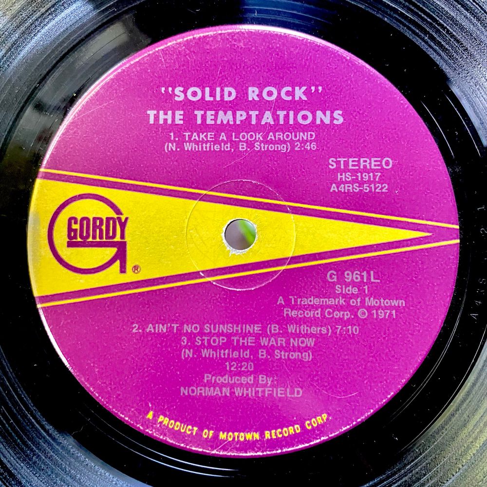 A close up image of a black vinyl record. The inner label is fuscia with yellow accent. Text reads “Solid Rock” The Temptations, with reflected light bringing a sense of depth and warmth. 