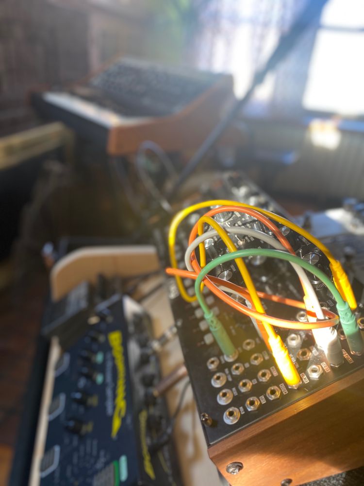A close-up shot of a modular synthesizer with colorful patch cables (yellow, orange, green) plugged into various inputs. In the soft-focus background, a Moog-style synthesizer rests on a stand in a warmly lit room with sunlight streaming through patterned curtains.