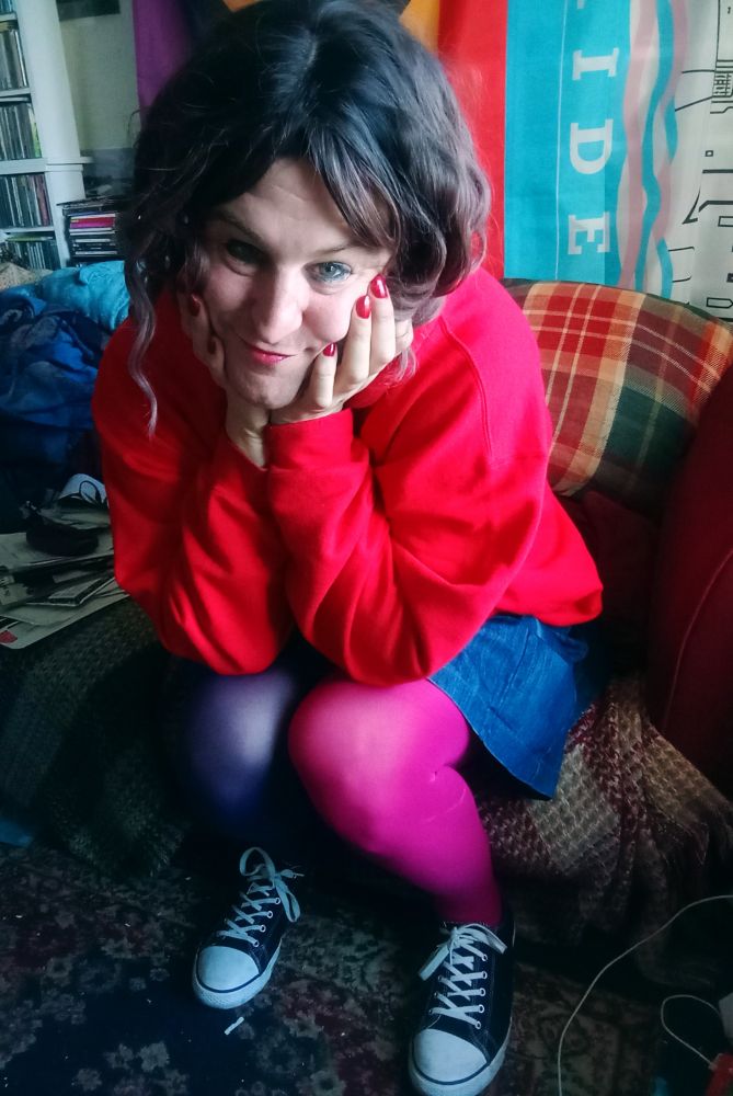 Pretty trans woman sitting on sofa in front of camera. She wears pink and purple tights, blue denim skirt and red hoodie. 