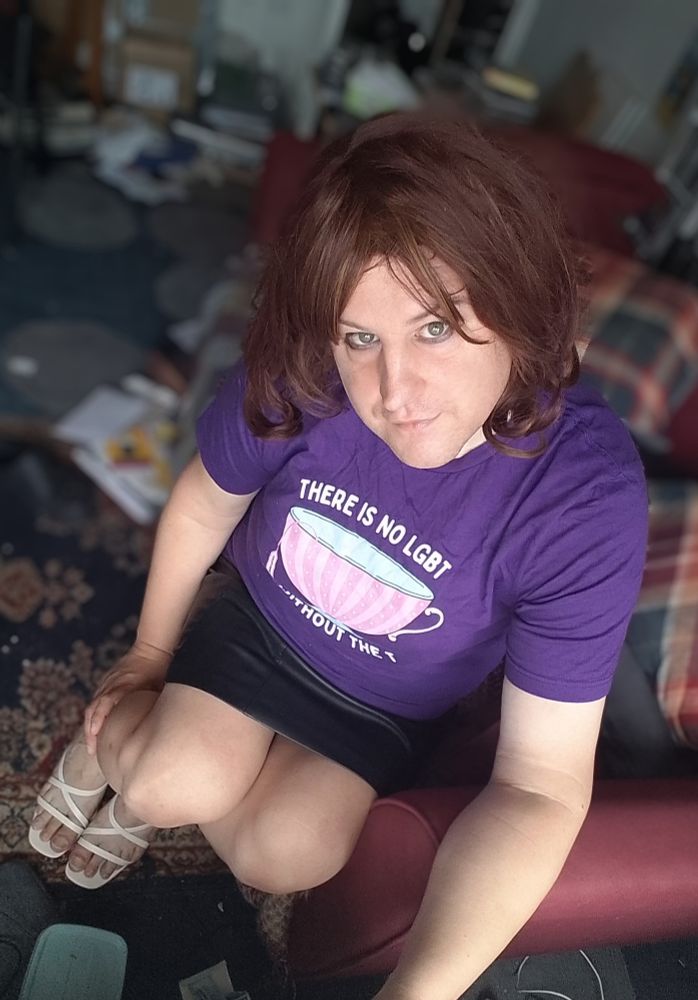 Pretty trans woman sitting on the sofa looking up at the camera. She has a black leather miniskirt, white heeled sandals and a purple t shirt on. Also possibly has wind.