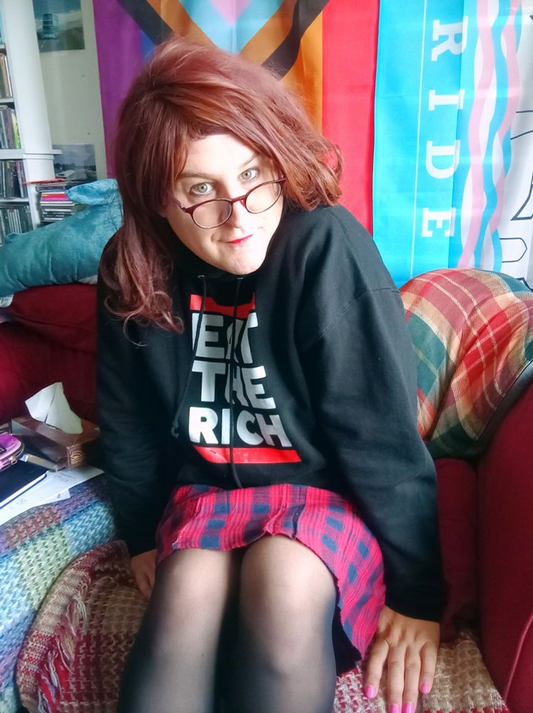 A pretty trans woman sits on a sofa, she wears a black hoodie, plaid red/black skirt and black tights. She has a pair of glasses sitting on edge of her nose. 