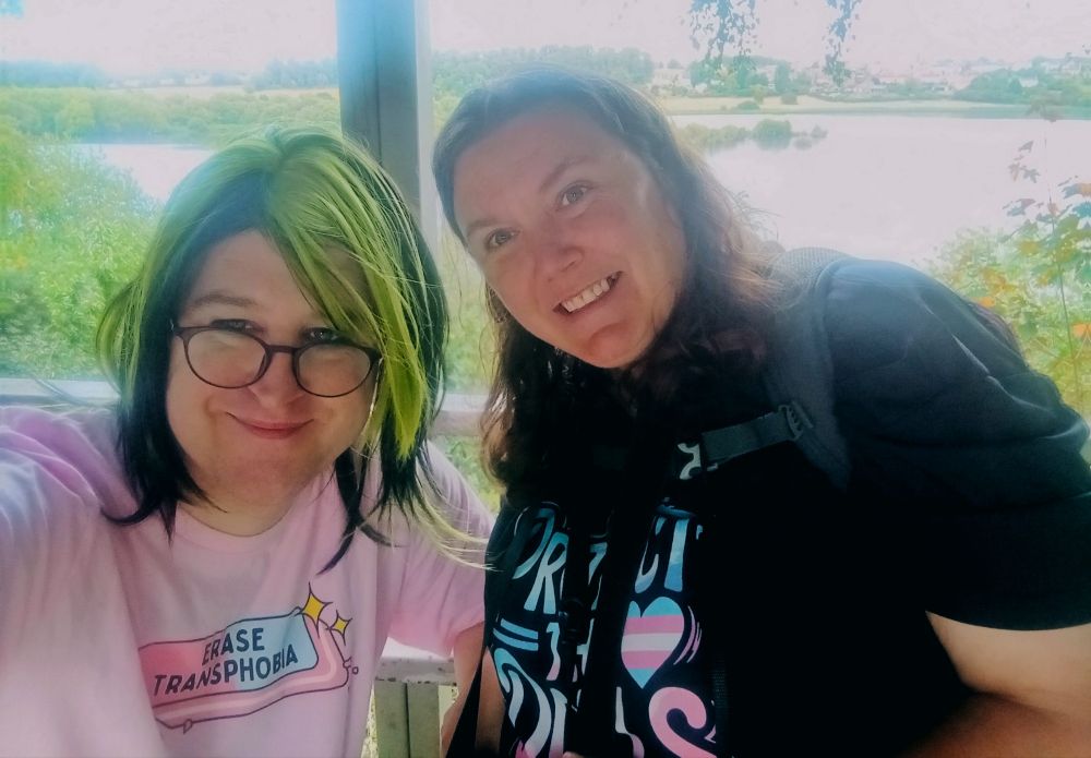 Pretty trans woman sitting a bird hide next to another pretty woman.  That pretty woman wears a black t shirt. 